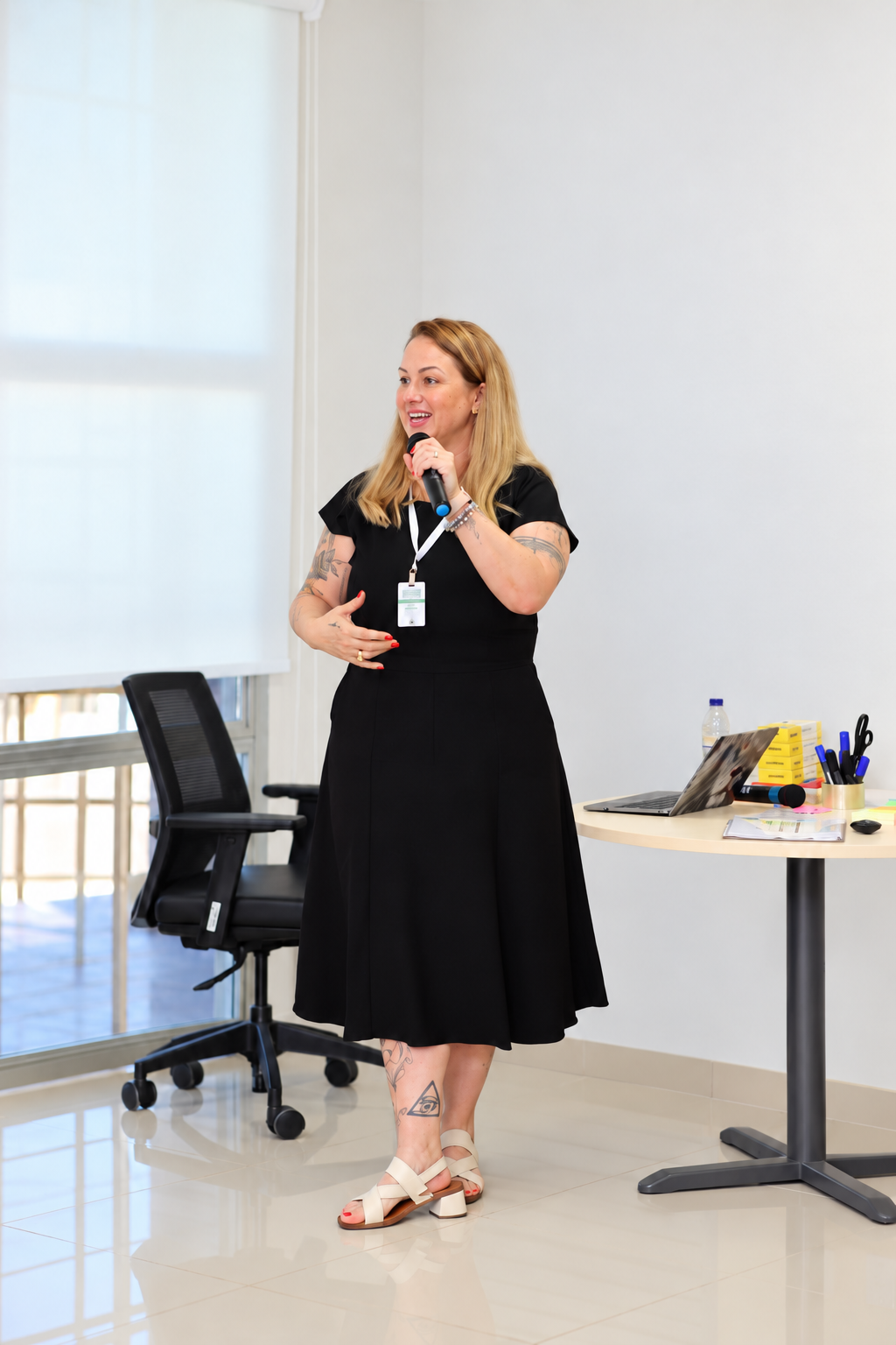 Camila Valentini palestrando com microfone em uma sala clara, em enquadramento de corpo inteiro.